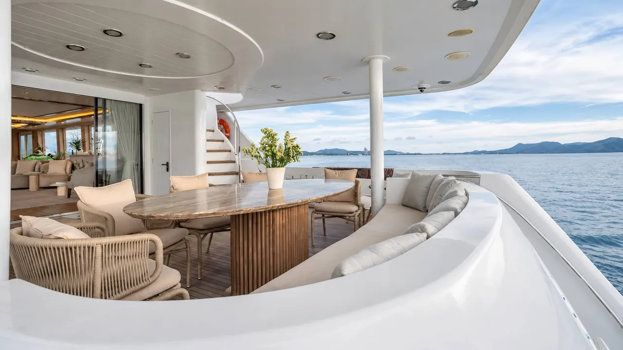 Shaded aft deck of Seabear with an oval dining table, rope chairs and a curved sofa overlooking the sea